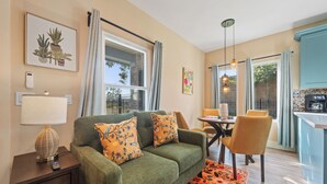 Een gezellige woonruimte met een bank, een eettafel met stoelen, een klein bijzettafeltje met een lamp, en een keuken met een aanrecht en hangende lampen.