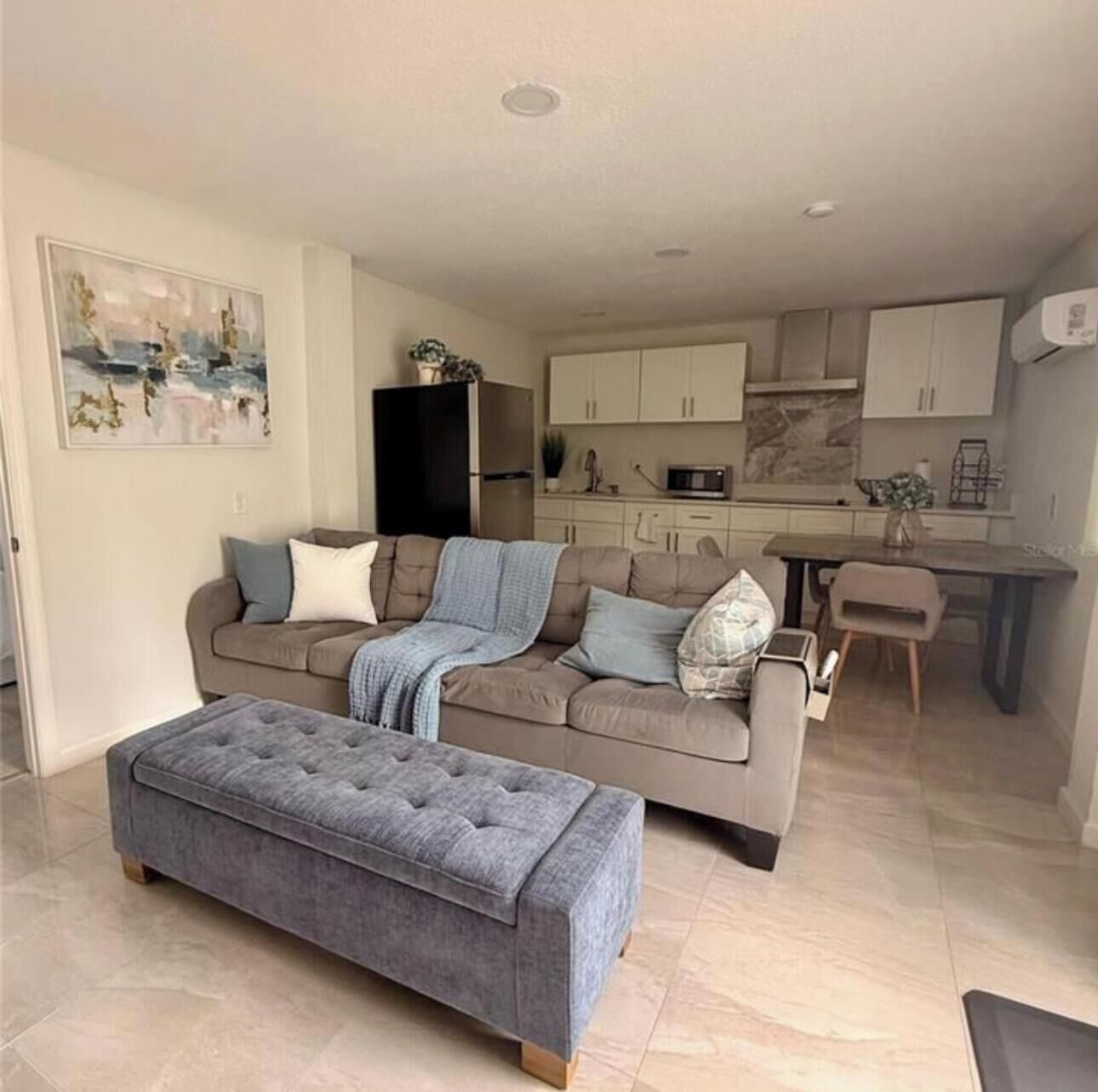 A modern living room with a grey sofa, a matching ottoman, and a dining area with a table and chairs.