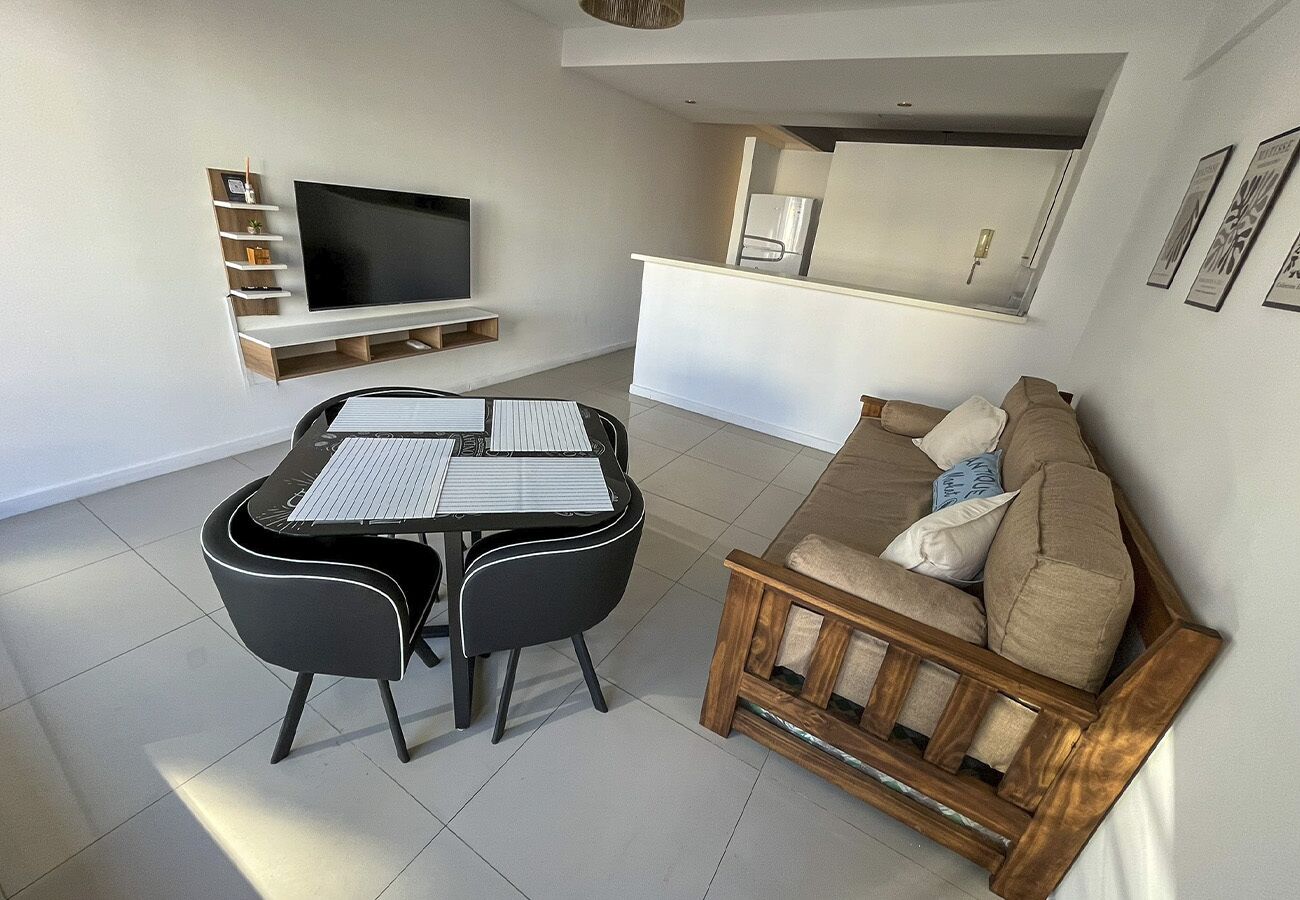 A modern living room with a dining table, black chairs, a wooden sofa, and a flat-screen TV mounted on the wall.