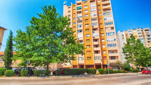 A multi-story residential building with balconies, a clear blue sky, and a street with parked cars and pedestrians. - Holiday apartment with air conditioning (Pula)