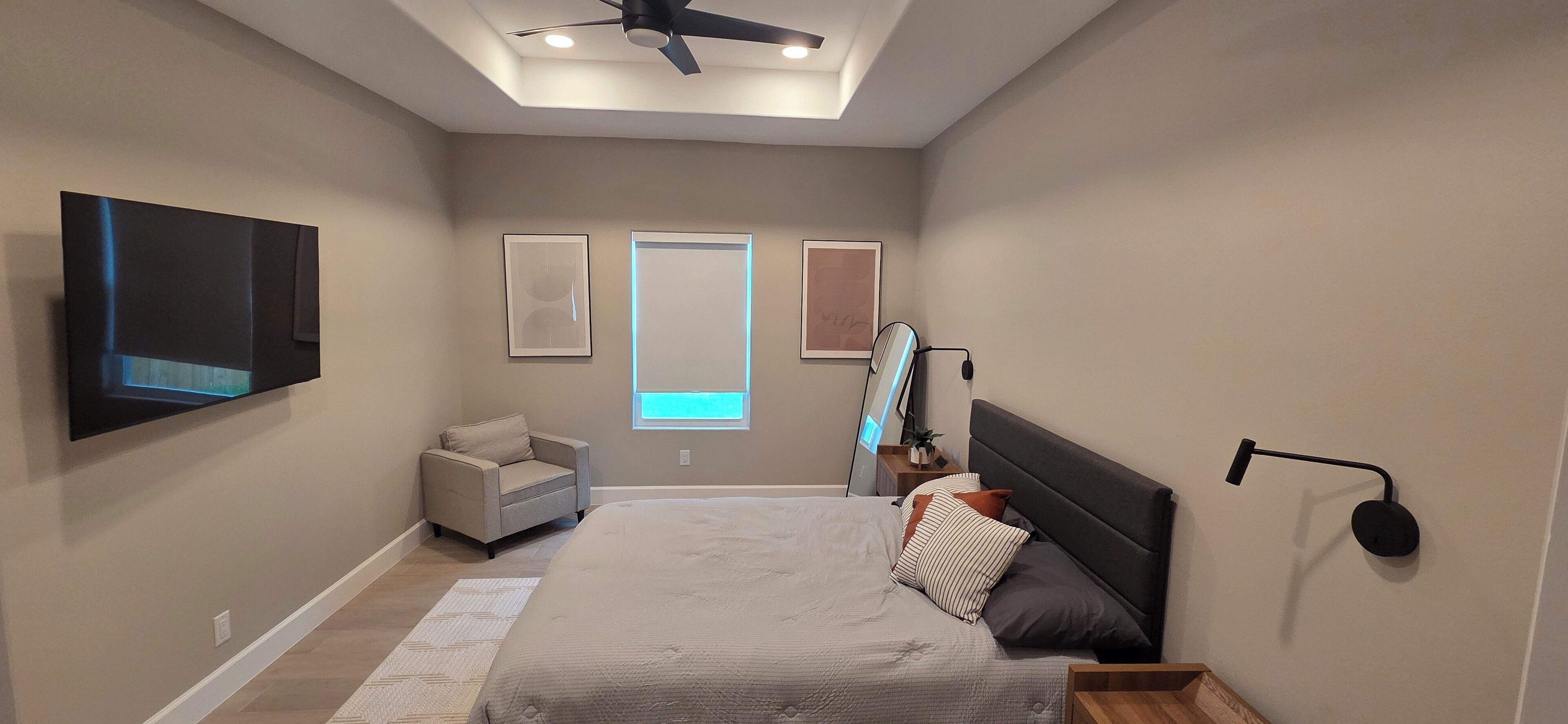 A bedroom with a bed, a TV mounted on the wall, a ceiling fan, and a window with daylight.