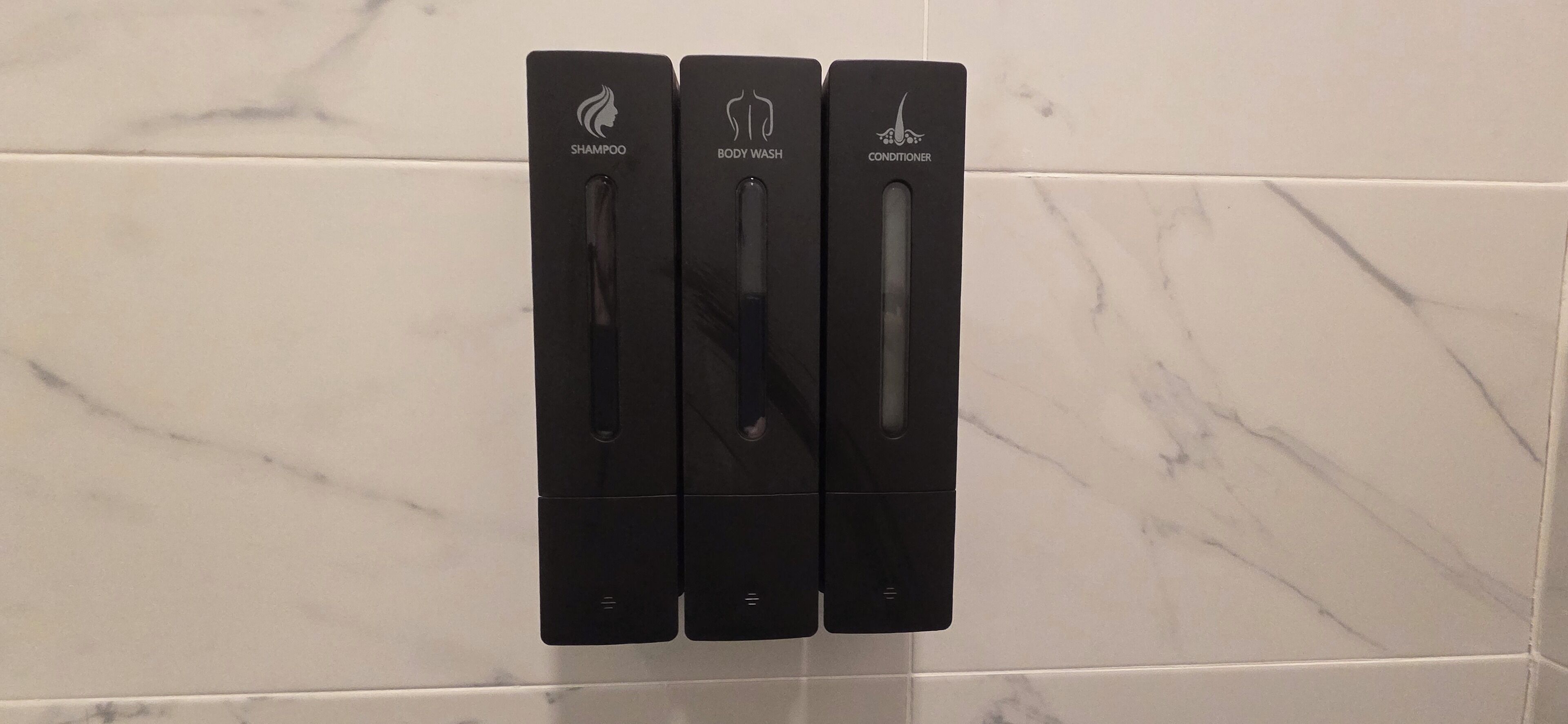 A set of black dispensers for shampoo, body wash, and conditioner mounted on a tiled wall.