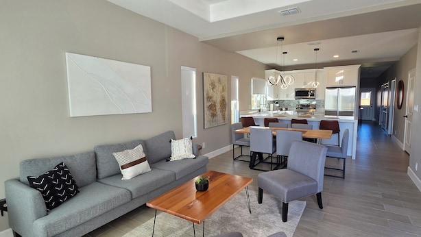 Un salon moderne avec un canapé gris, une table basse en bois et un fauteuil gris. On y trouve également un coin cuisine avec une table à manger et des chaises, ainsi qu'un couloir menant à une autre pièce.