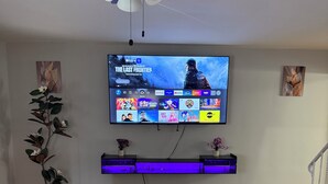 Una sala de estar con televisor de pantalla plana montado en la pared, piso de madera, ventilador de techo y una planta decorativa.
