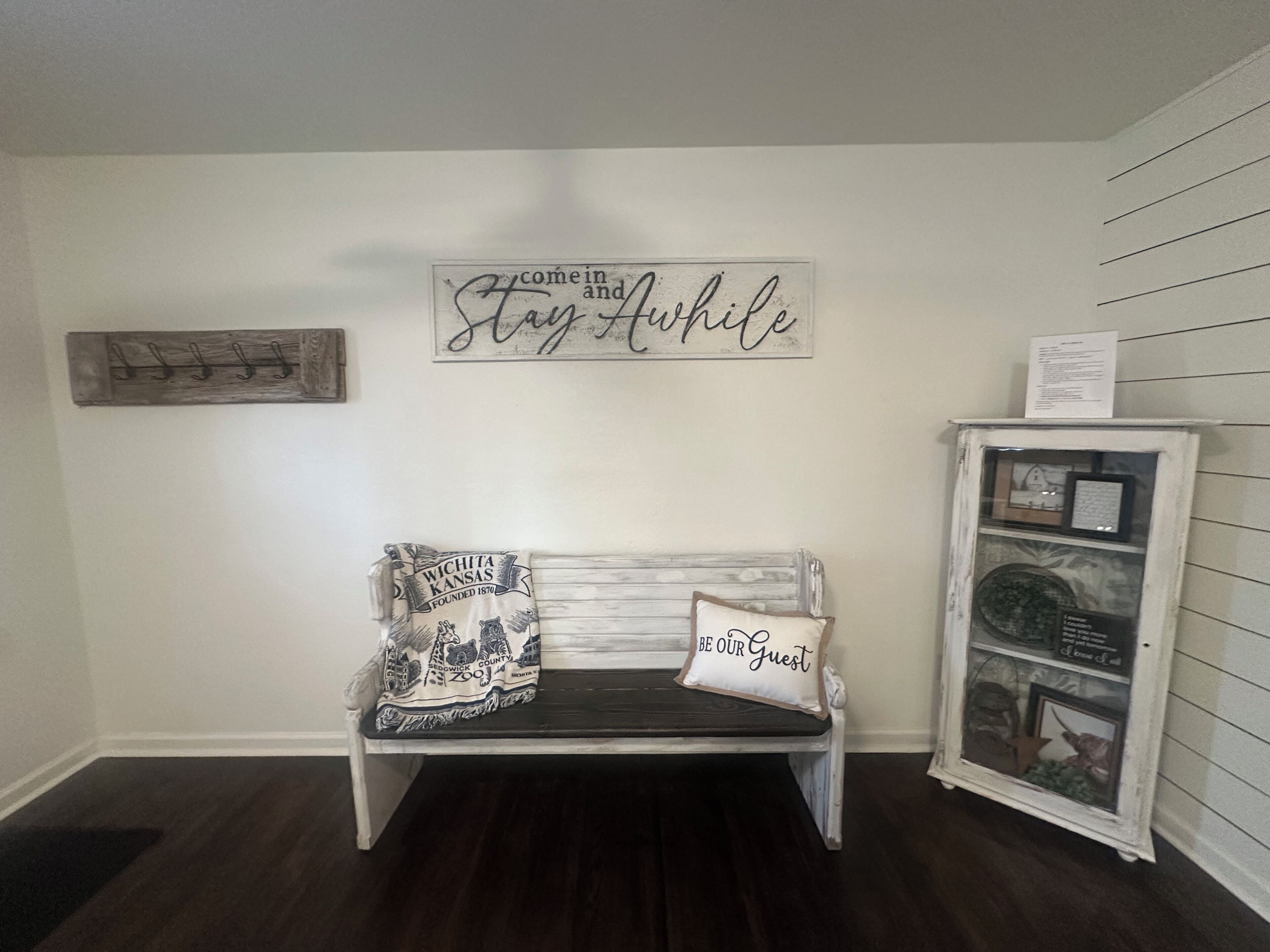 A room with a white bench, a sign saying "Come in and Stay awhile", and a cabinet with a glass door.