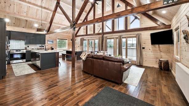A spacious living area with a wooden ceiling, a brown leather sofa, and a kitchen with modern appliances. - New Rangeley Mountain Home with Snowmobile Trail Access | Sleeps 9 (Rangeley)