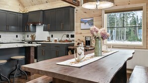 A modern kitchen with dark cabinets, a large island, and a view of the forest through a window. - New Rangeley Mountain Home with Snowmobile Trail Access | Sleeps 9 (Rangeley)
