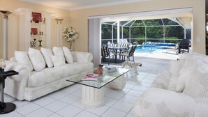 En rummelig stue med en hvid sofa, et glasbord og udsigt til en baghave med swimmingpool og udendørs møbler.