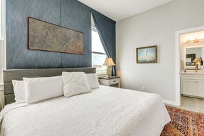 A hotel room with a large bed, a nightstand, a lamp, a painting, and a view of the city. - Rooftop Ocean View + Pool + Golf Cart (Port Aransas)