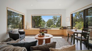 A modern living room with a sofa, a round wooden table, and a dining area with chairs. Large windows offer a view of trees and a clear sky. - Tranquility Base - 1 Bedroom Unit - Beachside (Coningham)