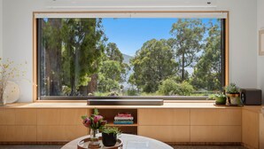 A room with a large window, a wooden bench, a round table with a book and a plant, and a bookshelf with books. - Tranquility Base - 1 Bedroom Unit - Beachside (Coningham)