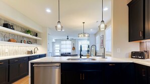A modern kitchen with a central island, stainless steel dishwasher, pendant lighting, and dark cabinetry.