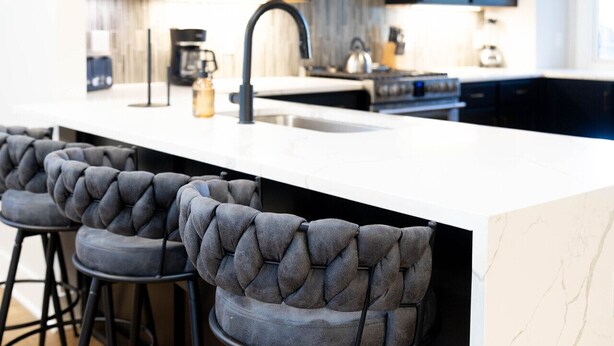 A modern kitchen with dark cabinets, a white marble countertop, and three tufted bar stools.