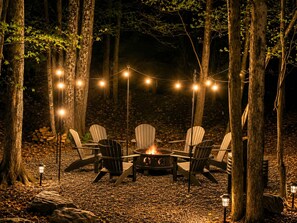 Ein Sitzbereich im Freien mit Adirondack-Stühlen, einem Feuerkorb und Lichterketten in einer Waldlandschaft.