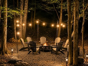 An outdoor seating area with Adirondack chairs, a fire pit, and string lights in a forest setting. - Mountain View Lodge by PMR, 8BR, Game Room, Hot Tub, Fire Pit, Sleeps 22 (Lake Harmony)