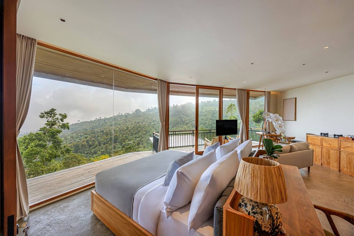 Ein modernes Schlafzimmer mit einem großen Bett, einem Holztisch und Blick auf eine bewaldete Landschaft.