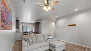 Ein modernes Wohnzimmer mit einem Ecksofa, einem Deckenventilator, Einbauleuchten und einem Essbereich im Hintergrund.