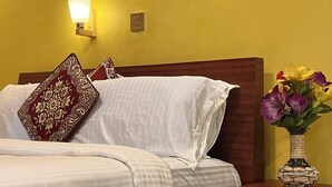Una cama bien hecha con sábanas blancas y almohadones estampados, una lámpara de noche y un florero con flores sobre una mesita de madera.