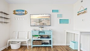 A room with a television, a white chair, a blue cabinet, and various wall decorations including a welcome sign and a happy place sign. - 1BR Oceanside Getaway: Beach, Pool & Views! (North Wildwood)