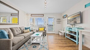 A modern living room with a grey sofa, a white coffee table, and a blue patterned rug. There is a television on the wall and a sign that reads "NORTH WILDO". - 1BR Oceanside Getaway: Beach, Pool & Views! (North Wildwood)