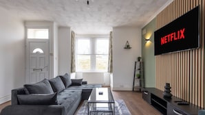 Uma sala de estar moderna com um sofá seccional, uma mesa de centro de vidro, uma TV de tela plana fixada em uma parede de madeira e uma janela com cortinas.