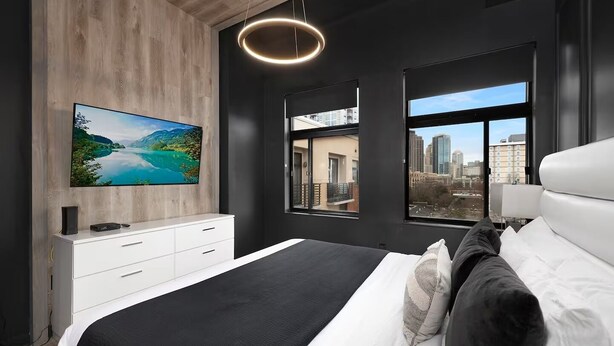 Un dormitorio moderno con una cama grande, un televisor montado en la pared, un aparador blanco y vistas a la ciudad a través de las ventanas.