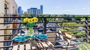 Un balcón con una mesa para dos, un florero con flores amarillas y una vista panorámica de la ciudad al fondo.