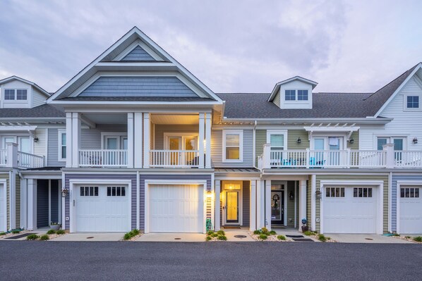 A row of two-story townhouses with attached garages, a covered front porch, and a small balcony on the second floor. - Luxury Shoal Harbor Retreat – Walk to Beach + Pool (Rehoboth Beach)