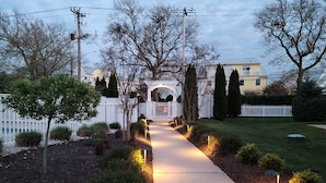 A pathway leading to a house with a white picket fence, surrounded by trees and shrubs. - Luxury Shoal Harbor Retreat – Walk to Beach + Pool (Rehoboth Beach)