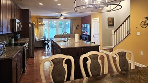 A modern kitchen with granite countertops, dark wood cabinets, a central island with seating, and a dining area with a chandelier. - Luxury Shoal Harbor Retreat – Walk to Beach + Pool (Rehoboth Beach)