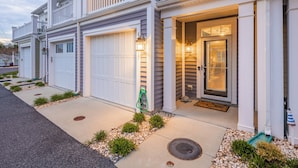 A two-story residential building with a garage, a front door with a glass panel, and a small porch area. - Luxury Shoal Harbor Retreat – Walk to Beach + Pool (Rehoboth Beach)