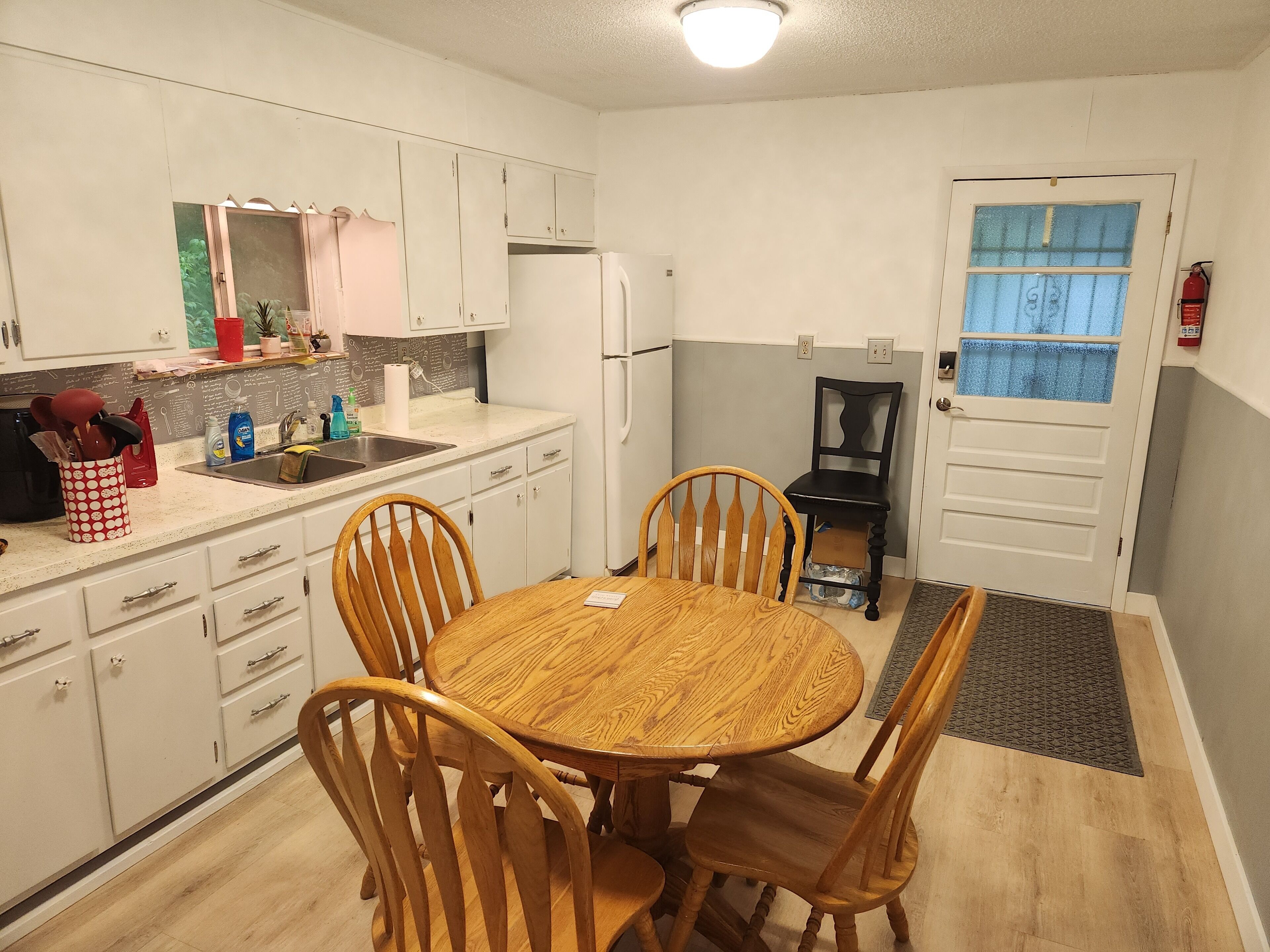 A compact kitchen with white cabinets, a sink, and a refrigerator. There is a dining table with four wooden chairs. A black chair is placed near the door.