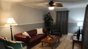 Una sala de estar con un sofá de cuero, un sillón verde, una mesa de centro de madera, un sillón negro, un ventilador de techo y una lámpara.