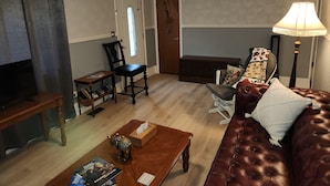 Una sala de estar con un sofá de cuero marrón, una mesa de centro de madera, una mesita auxiliar pequeña, una estantería y una lámpara.