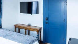 A bedroom with a bed, a wooden bedside table, a blue door, and a mounted flat-screen TV. - Van Wormer Room 201, Hyland Hotel (Palmer)