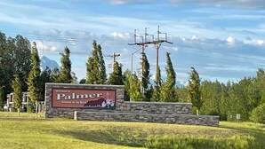 A welcome sign for Palmer, Alaska, with a stone wall and a view of trees and power lines in the background. - Bugge Room 203, Hyland Hotel (Palmer)
