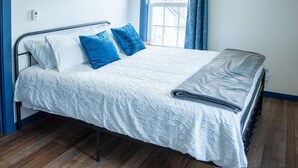 A neatly made bed with a white bedspread and blue pillows, a grey blanket, and a window with blue curtains. - Werner Room 204, Hyland Hotel (Palmer)