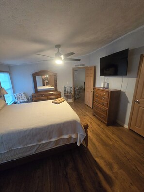 Un dormitorio con cama, cómoda de madera, televisor montado en la pared, ventilador de techo y puerta que da a otra habitación.