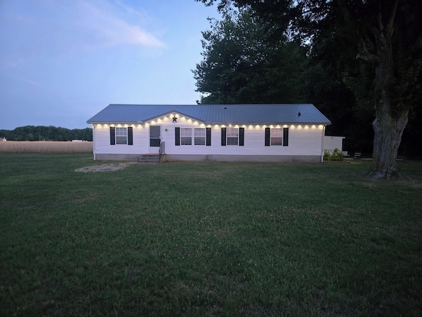 A white building with a metal roof, a front door, and a staircase leading up to it. There are lights along the roofline and a tree to the right.