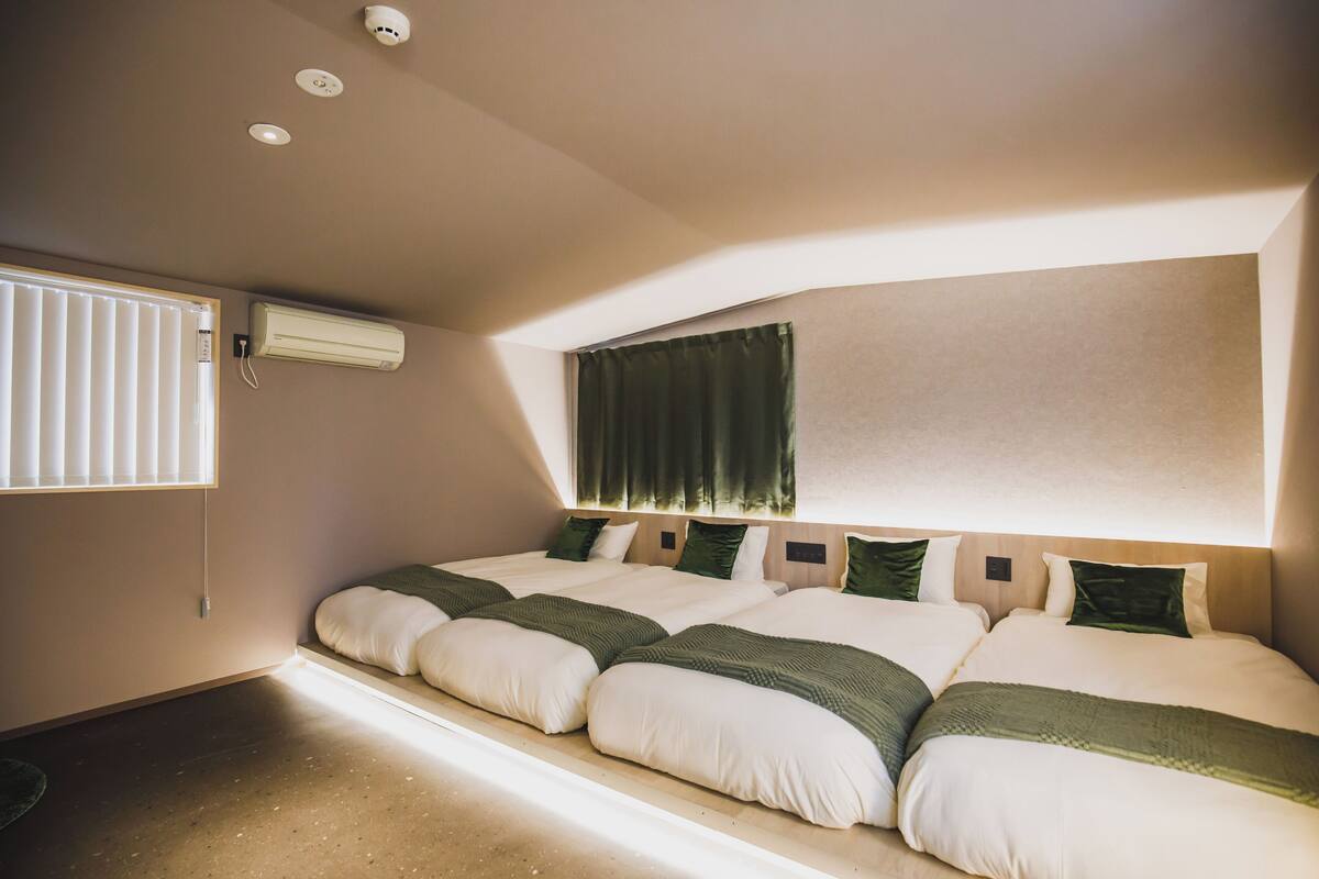 A hotel room with a large bed, a window with blinds, an air conditioning unit, and a ceiling-mounted security camera.