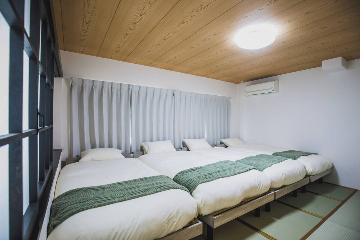 A hotel room with a wooden ceiling, a large window with curtains, a bed with a green blanket, and a tatami mat floor.