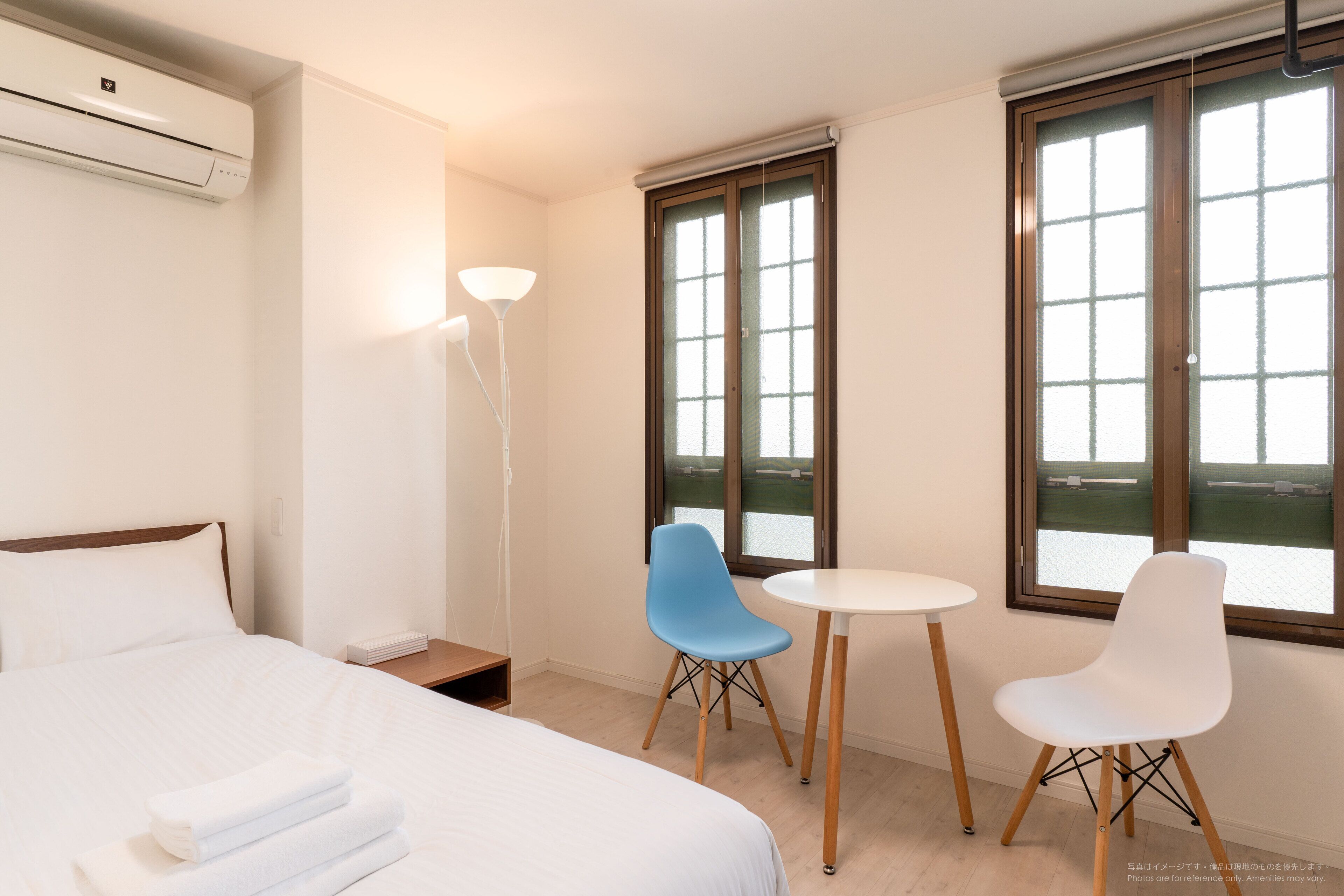 Une chambre d’hôtel avec un lit, deux chaises, une table, une lampe et de grandes fenêtres.