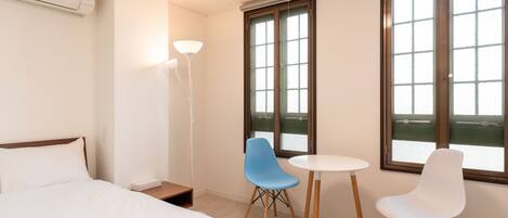 Une chambre d’hôtel avec un lit, deux chaises, une table, une lampe et de grandes fenêtres.