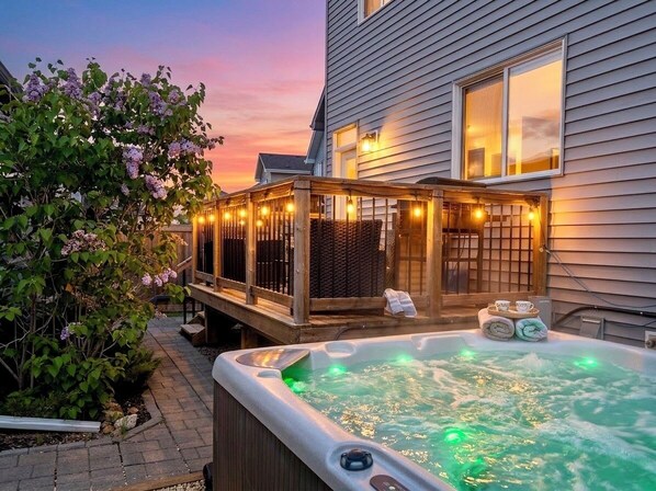 A hot tub and deck area with string lights, a wooden fence, and a house in the background.