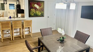 A modern kitchen with a white countertop and wooden chairs. There is a dining table with a woven tablecloth and a vase of flowers.
