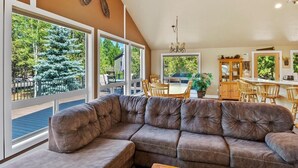 A living room with a large sectional sofa, a wooden coffee table, and a dining area with a table and chairs. There is a large window providing a view of trees and a deck outside. - Spacious 6-bedroom house in Sunriver perfect for families/groups (Sunriver)