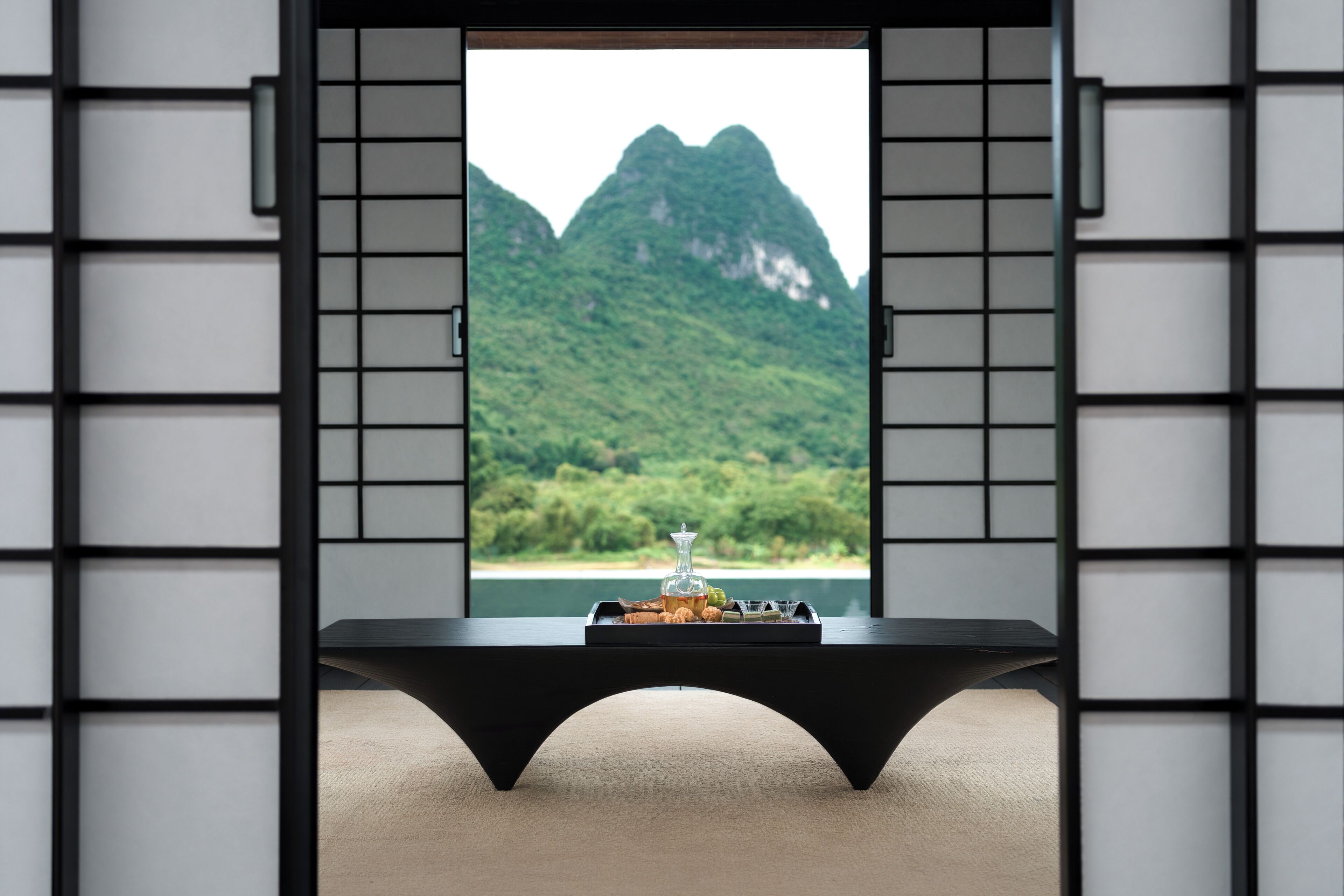 Una habitación con vista a montañas verdes a través de una ventana, una mesa negra con una bandeja de aperitivos y una silla negra.