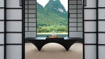 Una habitación con vista a montañas verdes a través de una ventana, una mesa negra con una bandeja de aperitivos y una silla negra.