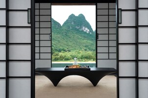 Una habitación con vista a montañas verdes a través de una ventana, una mesa negra con una bandeja de aperitivos y una silla negra.