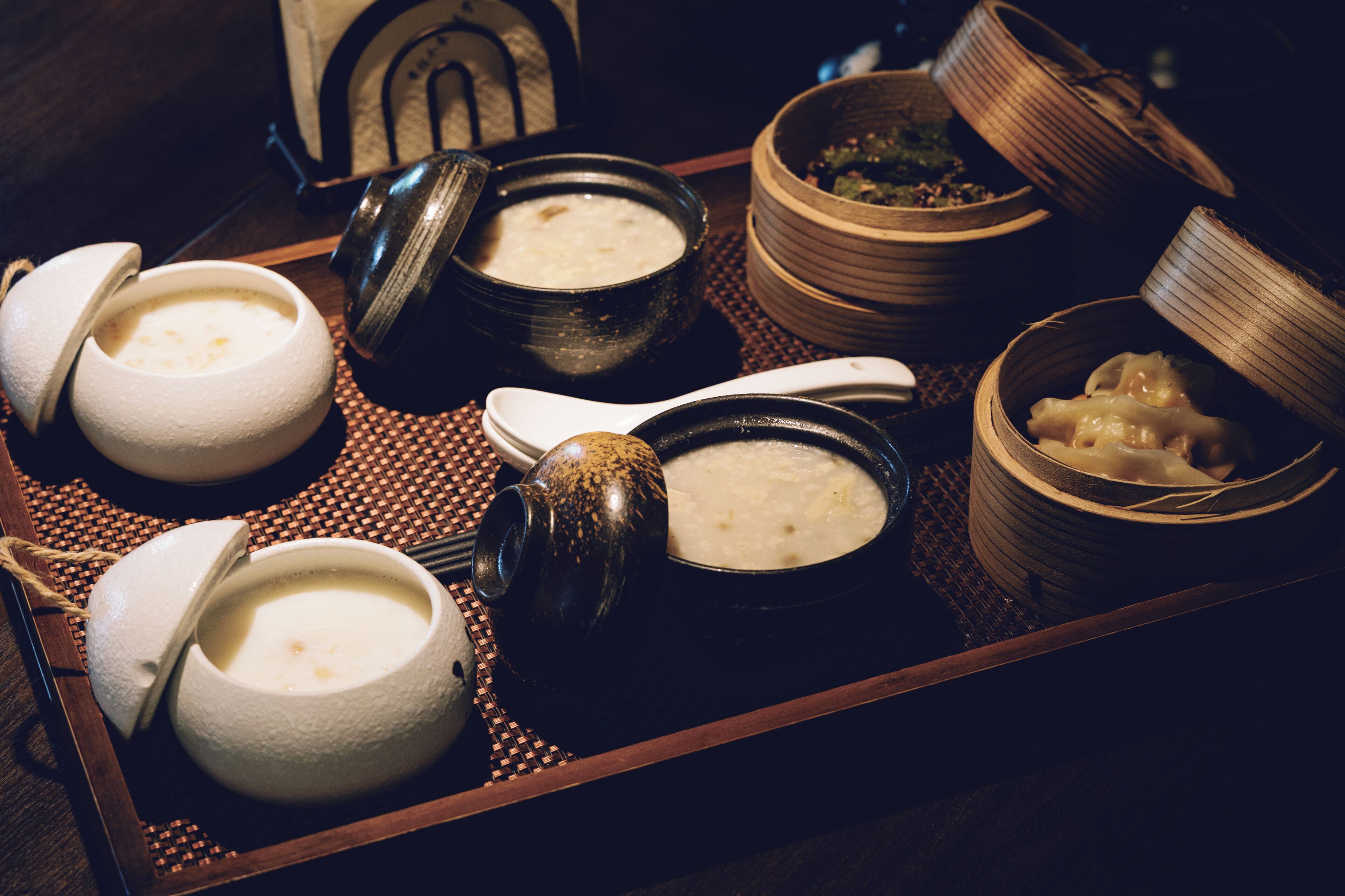 A tray with various bowls of food, including soup and dumplings, on a woven mat.